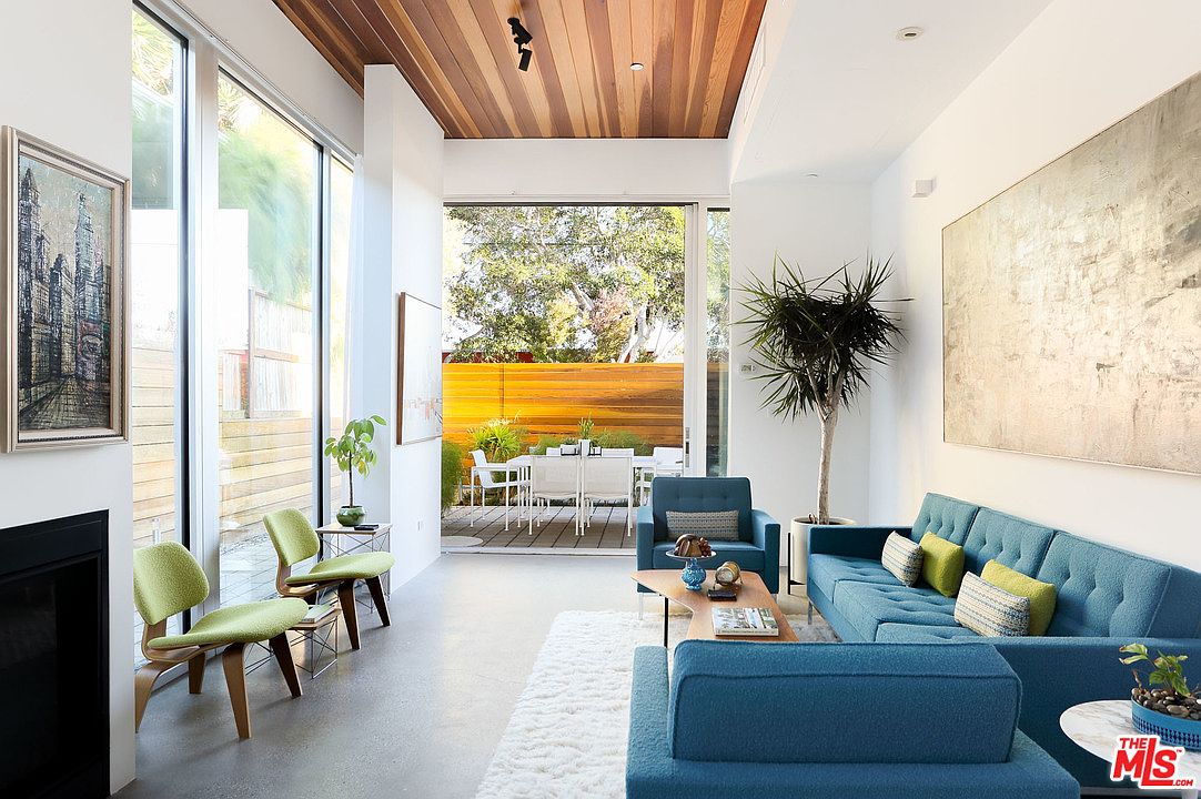 Living room with wood-beamed ceiling, retractable glass doors open to the patio, blue mid-century furniture, and polished concrete floor